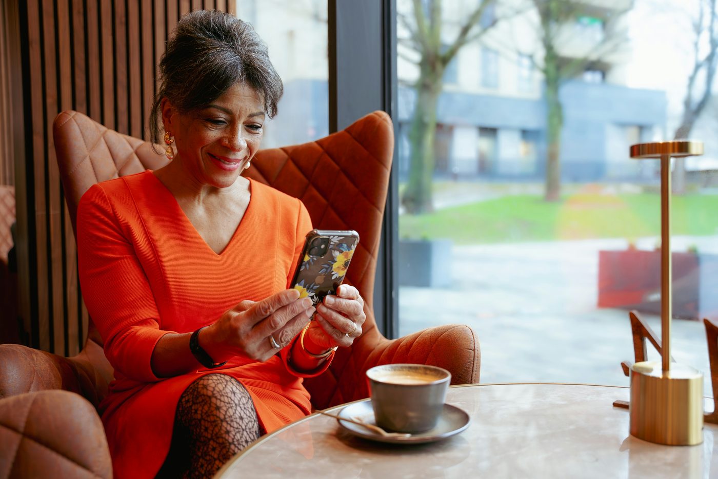An older woman sitting comfortably with her phone in a café, smiling at what she sees.