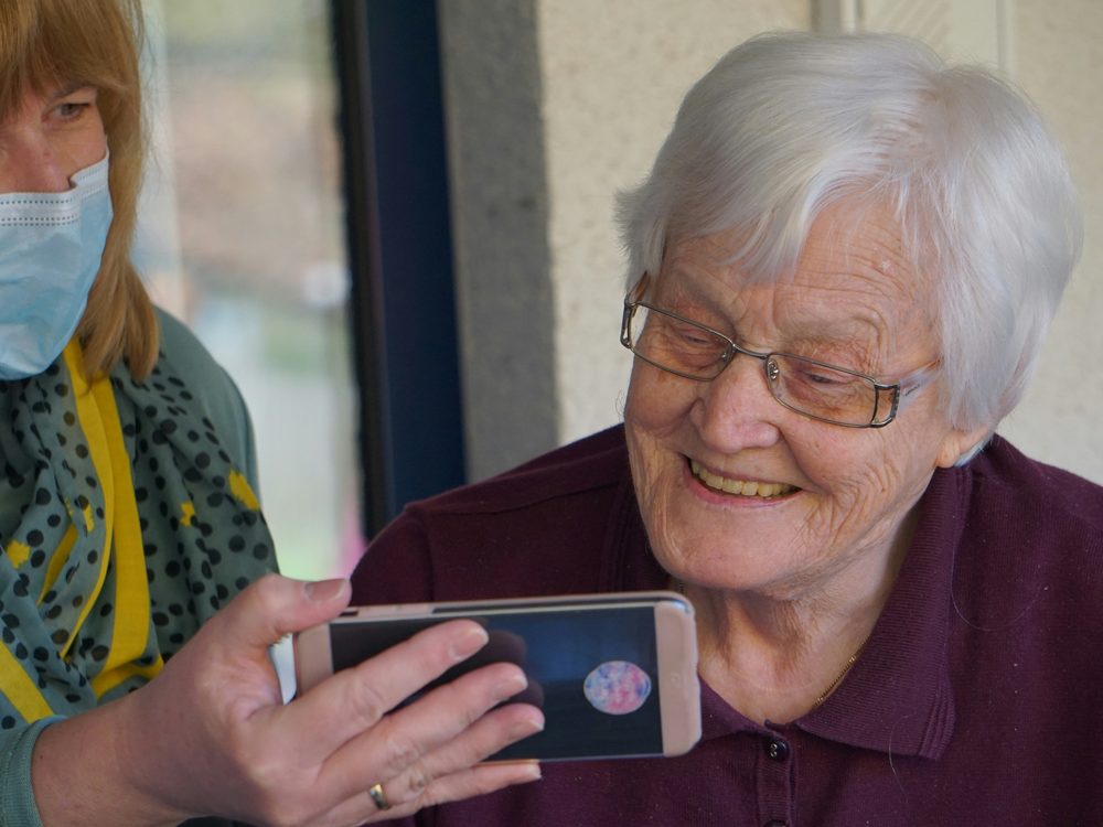 A care worker and an older woman laughing together over a phone screen.
