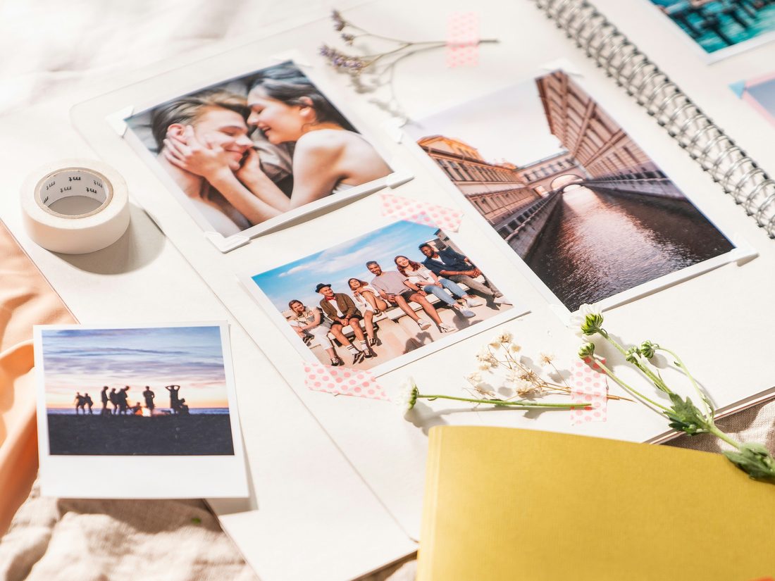 Printed photographs spread across a linen cloth beside an open album, taped down, with dried flowers among them.
