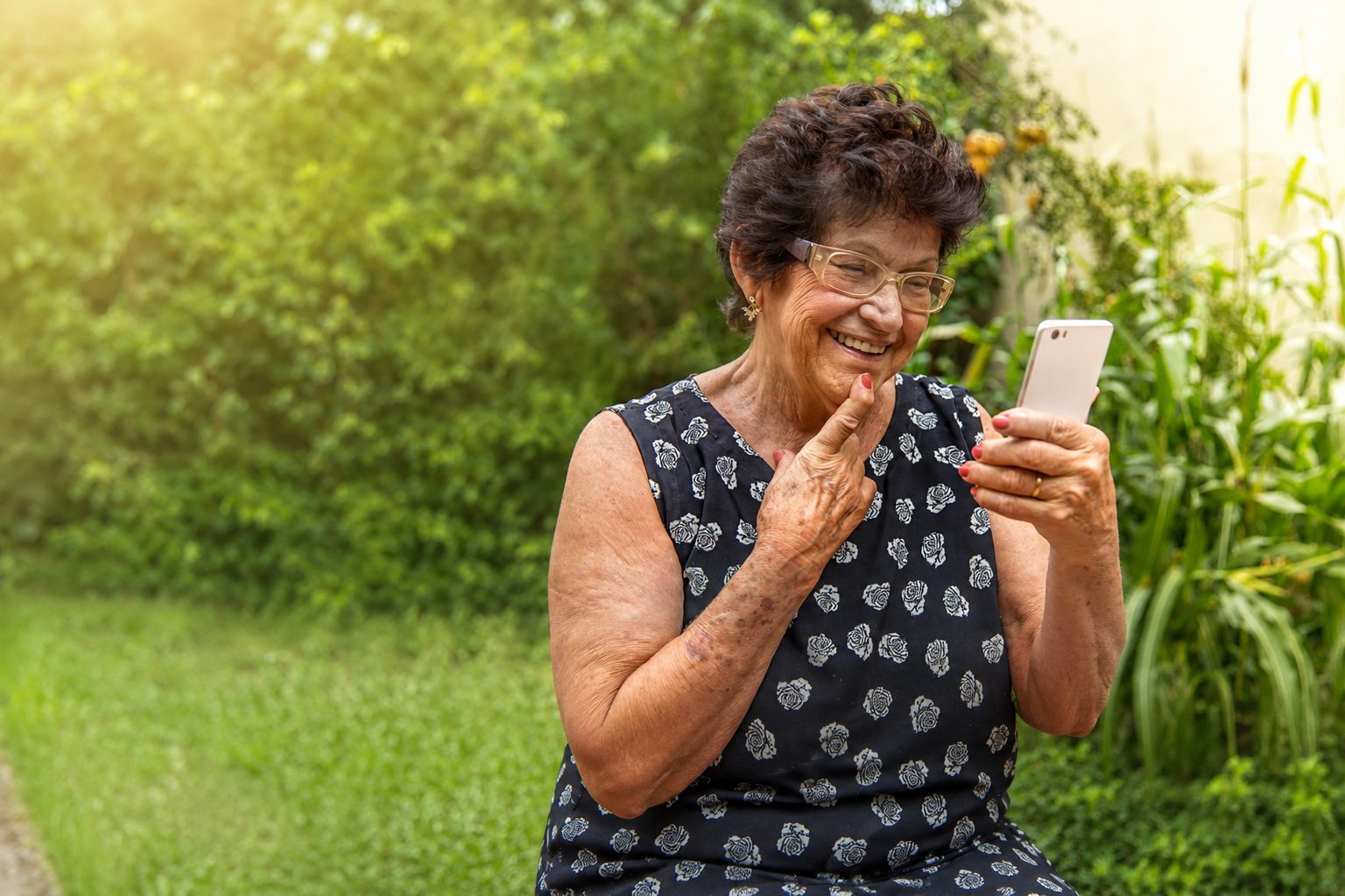 An older woman in a garden, hand at her chin, laughing at something playing on her phone.
