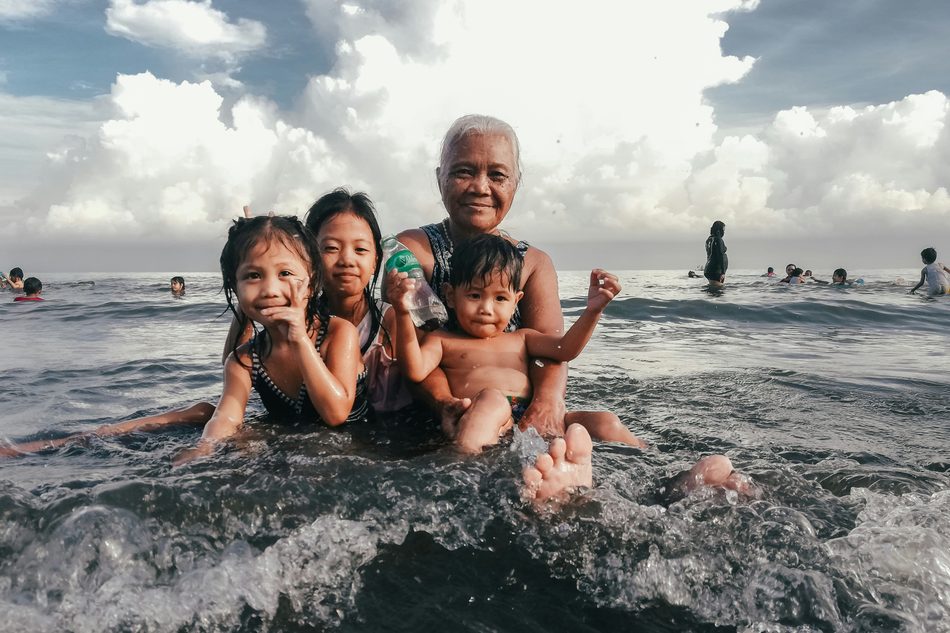 A grandmother in the sea with three grandchildren, all sitting in the shallows.